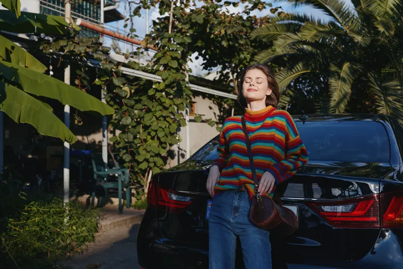 Woman leaning on black car trunk in tropical outdoor setting, wearing colorful striped sweater and jeans, smiling with eyes closed, casual purse and relaxed travel lifestyle vibe.