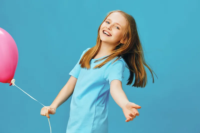 Cheerful girl with long hair holding a pink balloon, wearing a light blue dress, smiling joyfully against a bright blue background, embodying happiness and childhood innocence