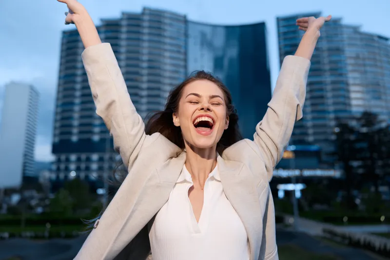 Joyful businesswoman celebrating success outdoors in urban setting, wearing stylish blazer, evokes confidence and positivity with vibrant city backdrop.
