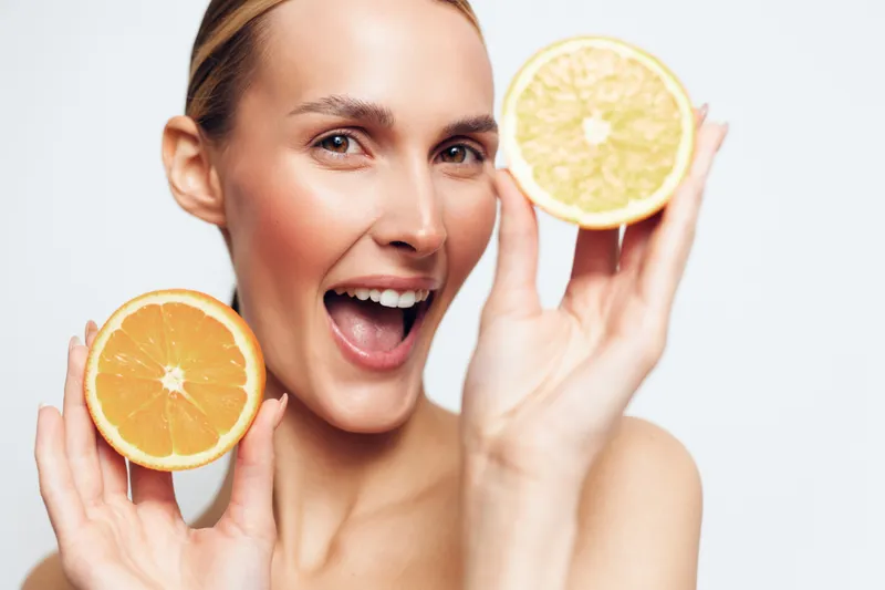 Happy woman holding two halves of citrus fruits near her face, smiling with open mouth, fresh natural skincare concept, bright studio lighting, clean white background