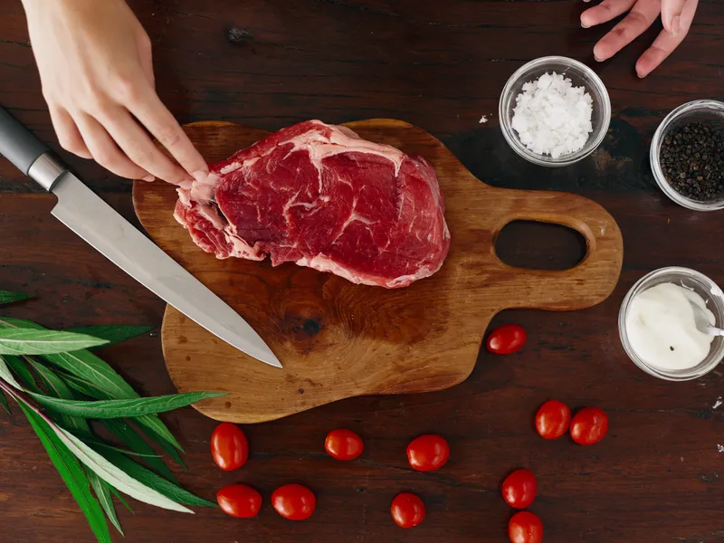 Женщина с ножом в руке нарезает свежее мясо стейка для жарки на кухне, на столе соль, перец и другие специи, красные помидоры черри и зелень, приготовление ужина в домашней кухне