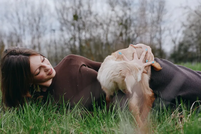 Woman lying on grass in a cosy sweater while a goat kid nudges her hand, capturing warmth, companionship, and rustic charm in an outdoor setting. Natural light creates a calm, lifestyle mood.