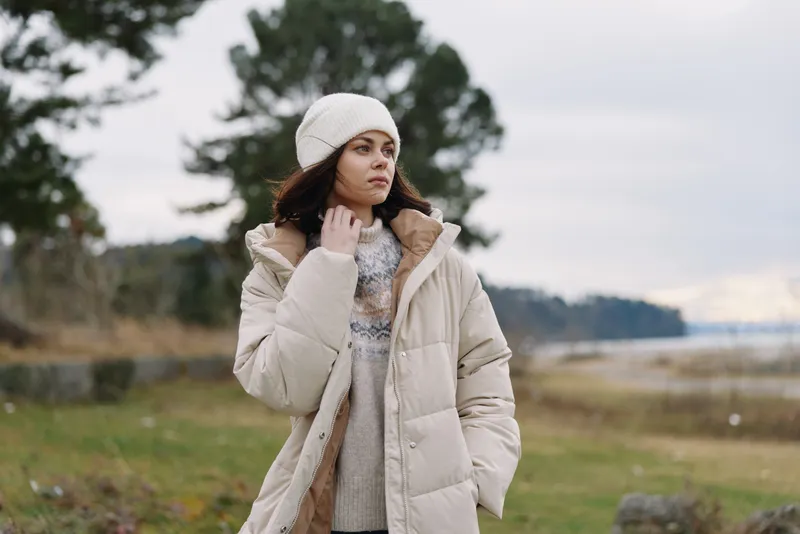 Winter scene with a thoughtful woman in a light beige coat and knit beanie, standing in an open countryside landscape, soft breeze, calm mood, natural textures and muted tones.