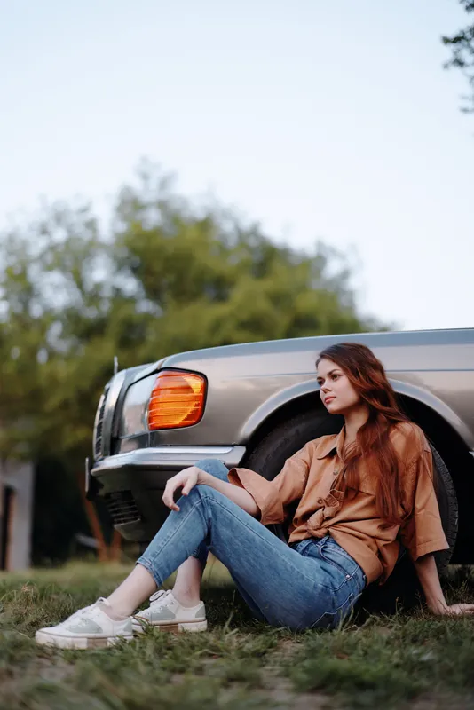 Young woman enjoying a quiet moment outdoors next to a vintage car, wearing casual attire with a relaxed expression, surrounded by nature in a serene setting.