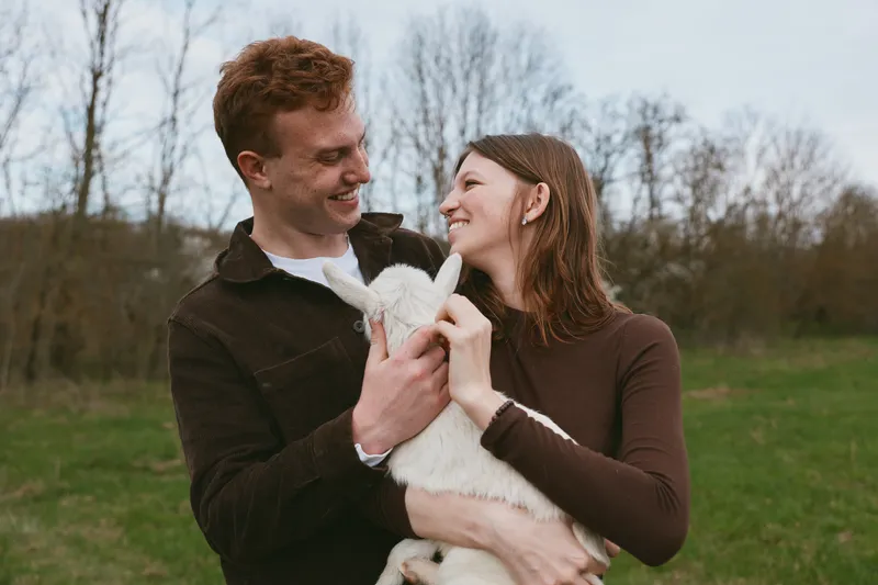Young couple holding a white baby goat outdoors in a green field during autumn, sharing a joyful moment together in casual brown clothing with natural light.