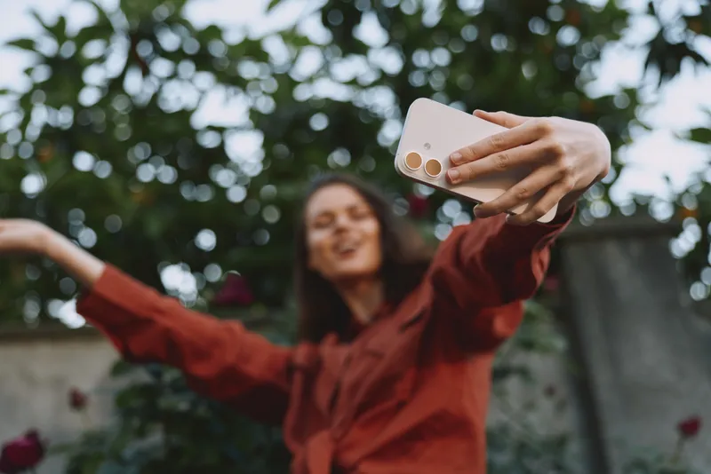 A smiling woman takes a selfie outdoors in casual attire, capturing a playful moment in a sunlit garden. Concept for everyday joy, social media, and personal branding. mood