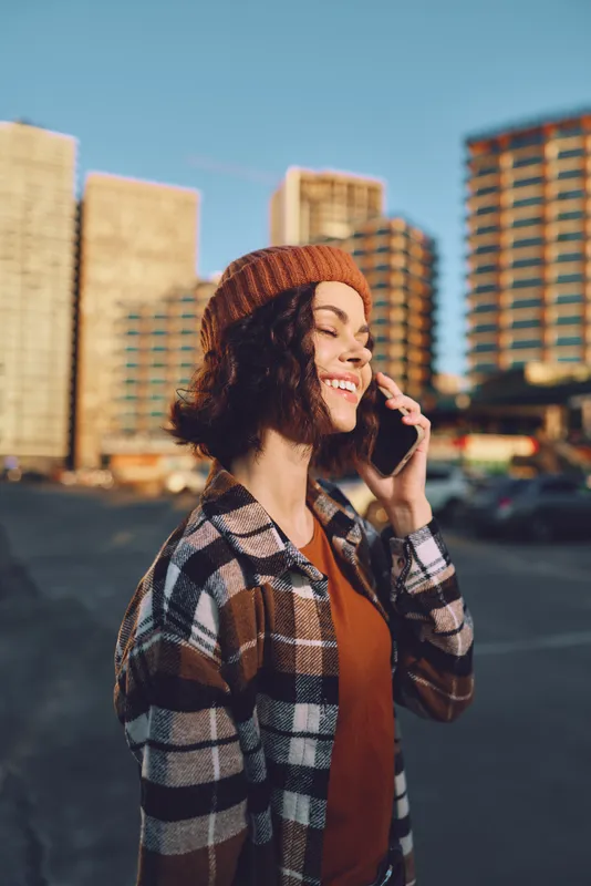 Woman on phone smiling in urban sunset, candid lifestyle portrait with beanie and plaid jacket. Golden hour glow, authenticity and emotional storytelling capture mindful living moment.