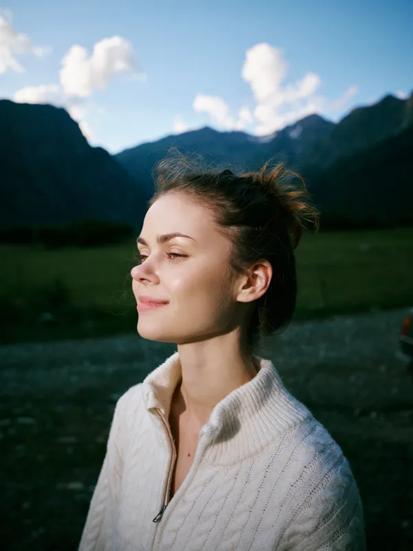 Woman in a cozy sweater stands in a calm valley with distant mountains, enjoying warm sunlight and fresh air, capturing a tranquil moment of outdoor life and nature