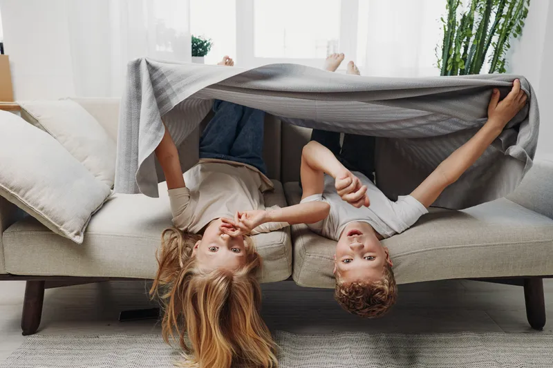 Two playful children lie upside down on a beige sofa covered with a gray blanket in a bright living room. The cozy scene shows family joy and childhood fun at home.