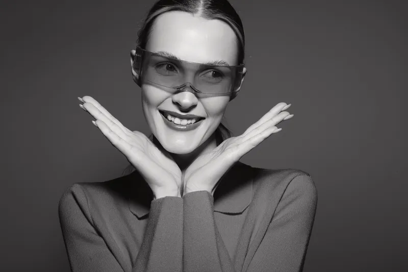 Young white woman with light skin wearing stylish sunglasses smiles happily with hands framing face. Studio portrait with casual clothing and modern eyewear concept.