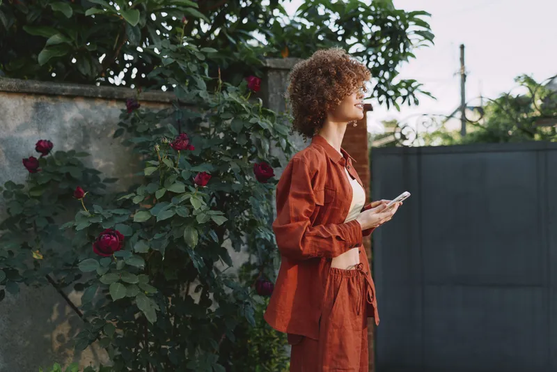 Curly haired woman in an orange outfit stands in a garden with roses, looking at a smartphone. Natural daylight highlights a stylish, relaxed lifestyle moment