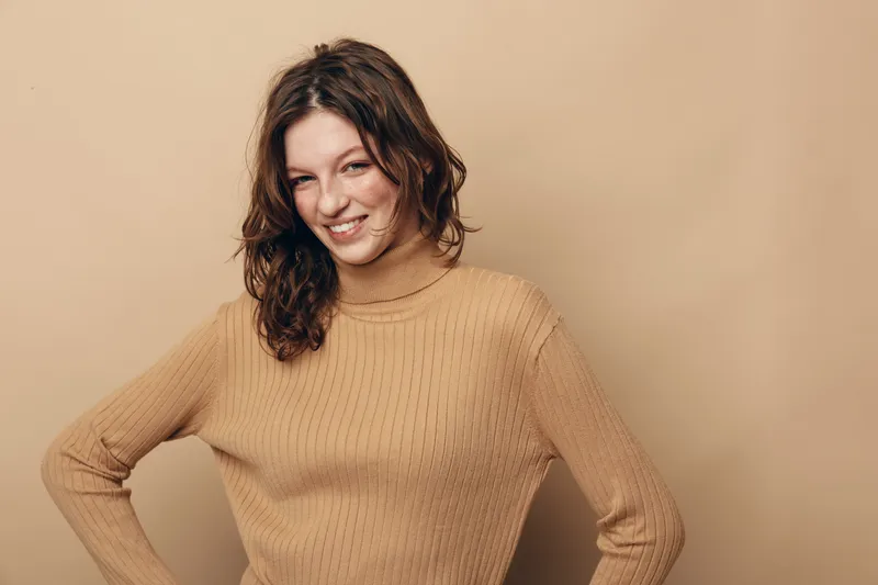 Smiling young woman with curly brown hair wearing beige knitted sweater stands against beige background. Portrait of confident female model with casual style and natural makeup in studio.