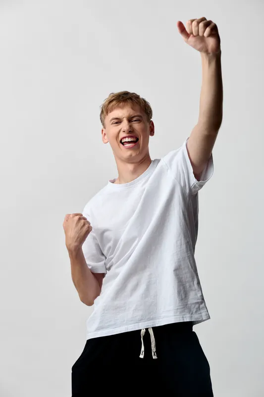 Celebration, victory, excitement and energy as a young man raises his fist in triumph, wearing a white t shirt and drawstring shorts against a plain backdrop, conveying motivation, achievement and