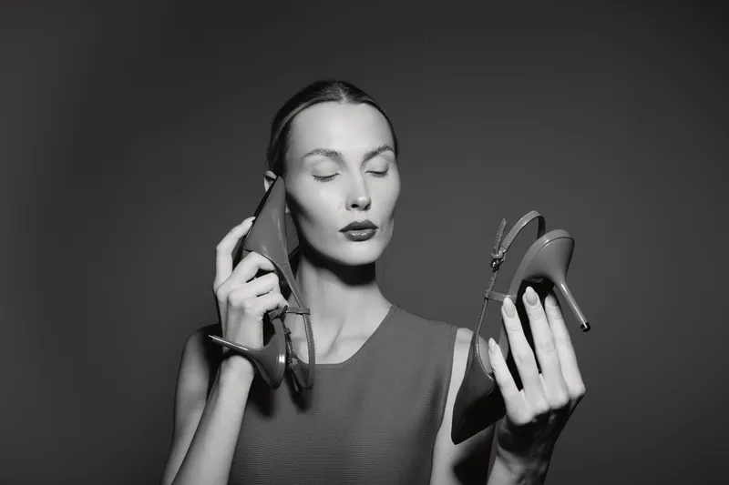 Young white woman with fair skin holding two high heel shoes, eyes closed in thoughtful expression, studio portrait with dark background.