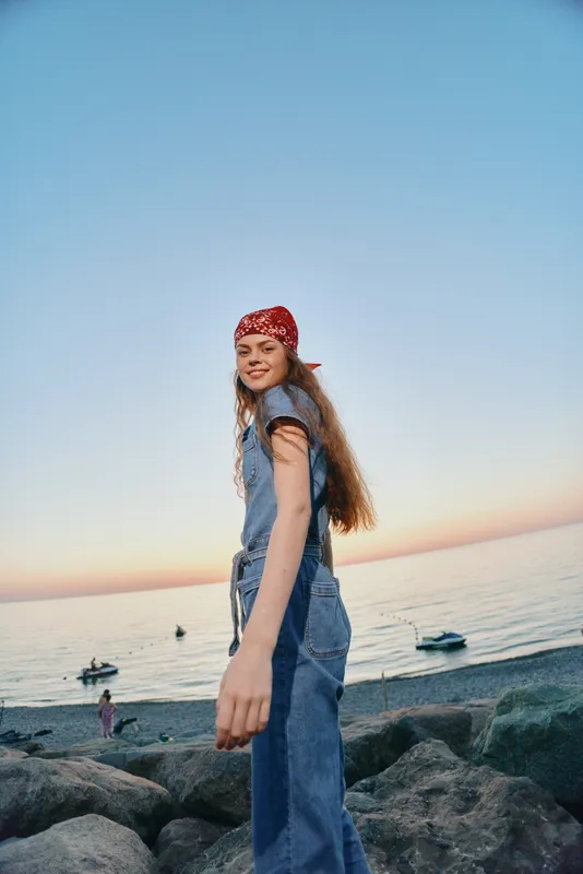 smiling woman wearing denim overalls and red bandana enjoys seaside sunset with calm ocean and boats in background, capturing relaxed and joyful mood