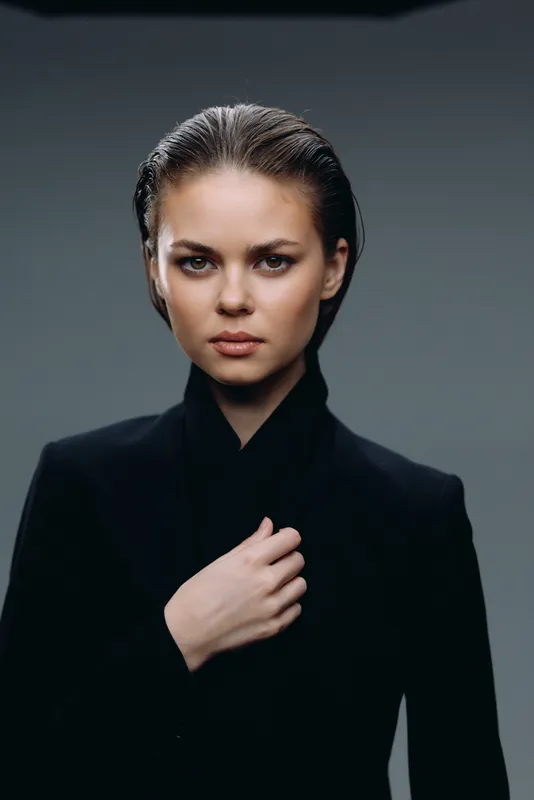 serious woman with slicked-back hair wearing black jacket with hand on chest against gray background