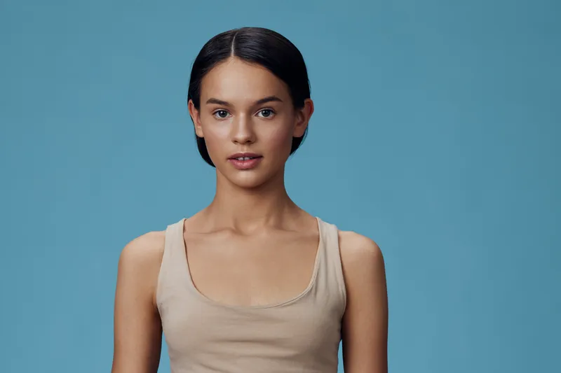 Young female model with long dark hair wearing a simple beige top against a soft blue background, exuding confidence and elegance