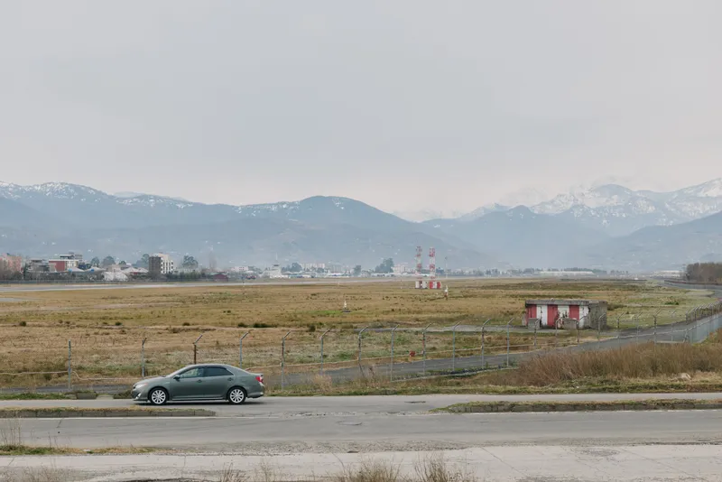 A grey car parked near an empty field with dry grass under an overcast sky. Mountains with snow caps are visible in the background, creating a calm and isolated landscape scene.