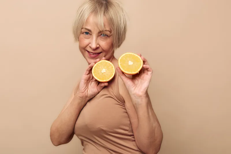 Smiling mature woman with short blonde hair holds two halves of fresh orange fruit near face on beige background. Concept of healthy lifestyle, vitamin intake, natural nutrition and wellness.