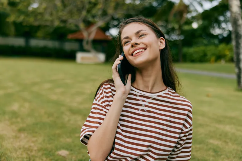 Счастливая женщина разговаривает по мобильному телефону, сидя на траве под пальмой в парке