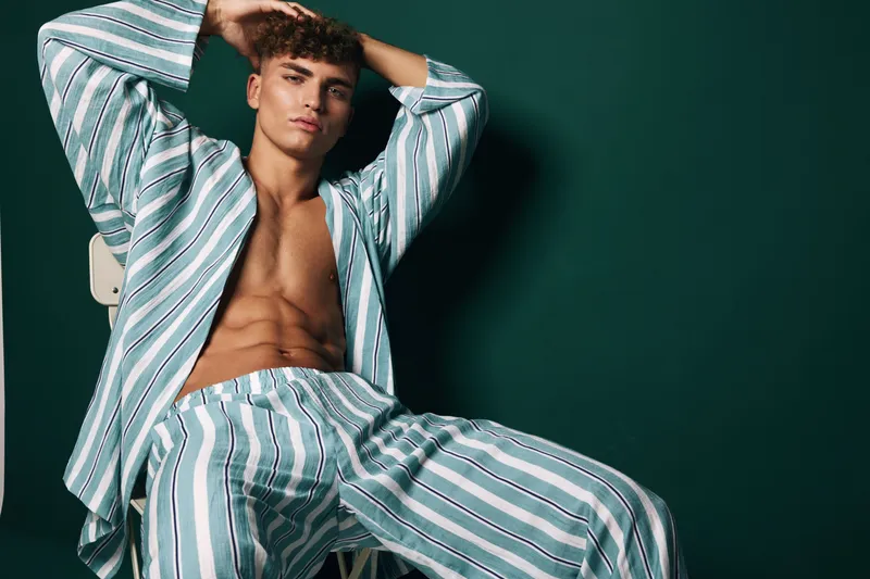 Young man in striped casual shirt and pants posing while sitting on chair with hand in hair relaxed against dark background. Fashion, model and lifestyle with grooming and fitness.