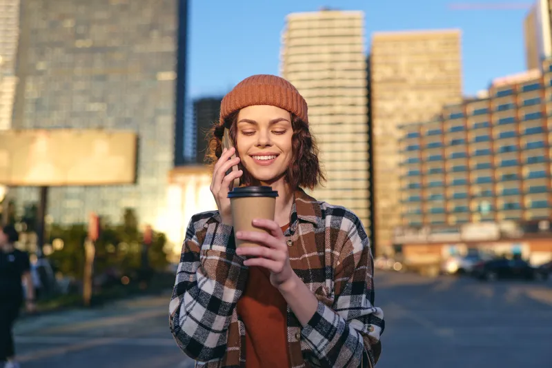 Woman holding coffee and phone on an urban street, smile and beanie in golden hour glow, candid lifestyle portrait emphasizing authenticity, mindful living and emotional storytelling.