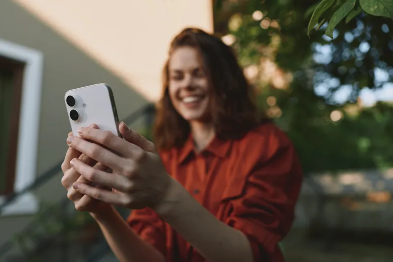 Young woman in a red shirt smiles at her phone while taking a selfie outdoors, capturing a bright casual lifestyle moment with natural light and greenery in the background