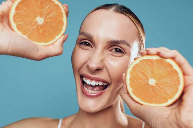 A joyful woman holds two orange halves against a bright blue backdrop, highlighting fresh fruit, a radiant smile, lively energy, and a healthy, vibrant lifestyle.