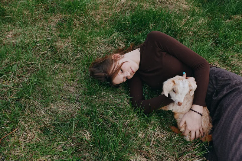 Young woman lies on grass holding a baby goat close in a relaxed outdoor lifestyle moment during springtime in casual clothing.