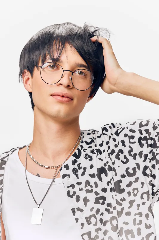 young man with round glasses wearing a patterned shirt and silver chains posing against a white background with a casual and modern style
