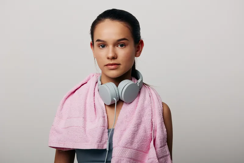 Woman in athletic wear with headphones and a pink towel draped over her shoulders, ready for a workout. Focused pose in a studio conveys fitness, sport and personal wellness.