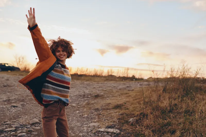 Curly hair person waving outdoors at sunset on a dirt road, warm golden sky and dry grass behind, lifestyle travel moment, casual clothing, soft natural light, carefree joy