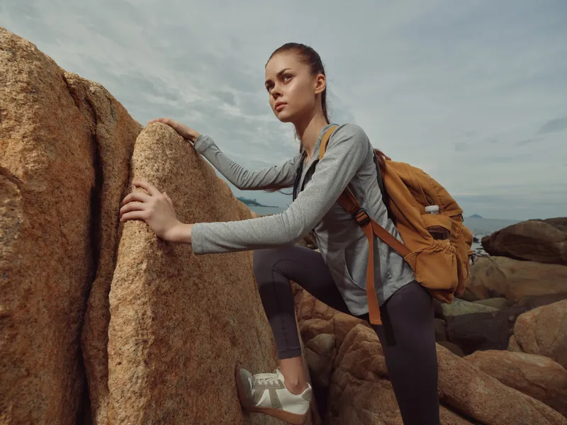 Young woman climbing rocks, wearing sportswear and a backpack, showcasing determination and adventure on a cloudy day by the sea