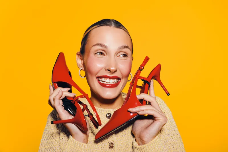 Happy woman with red lipstick holds red high heel shoes near face on bright yellow background. Concept of fashion, style, shopping, and joyful emotions.