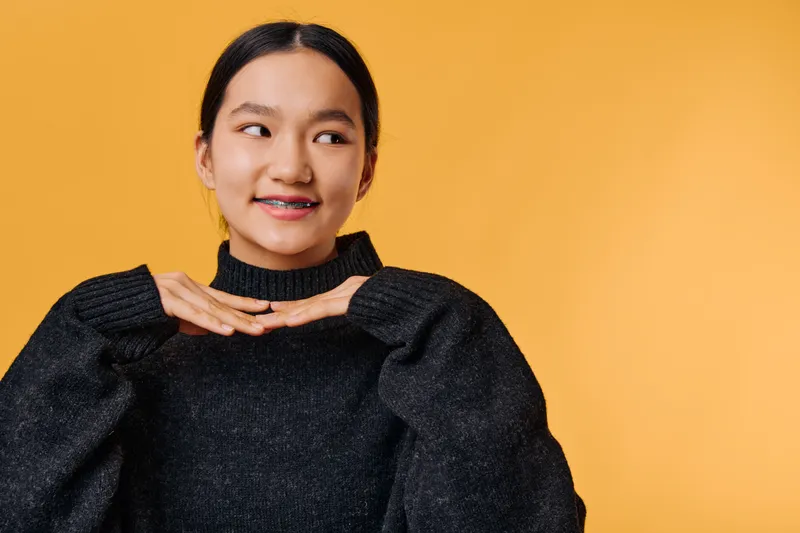Young woman wearing black sweater with braces smiling and posing with hands under chin against orange background. Portrait of cheerful female person with happy expression for lifestyle.