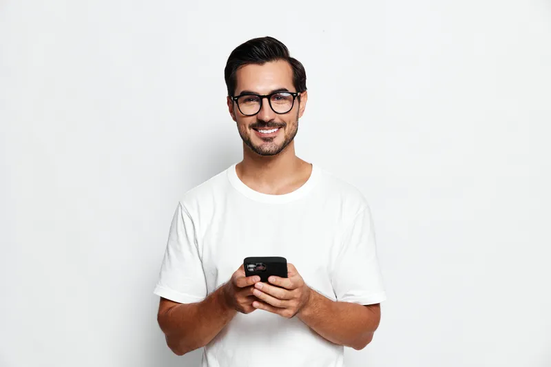 Smiling man wearing glasses holds smartphone in hand, staring at camera in casual white t-shirt against plain solid background. Lifestyle concept.