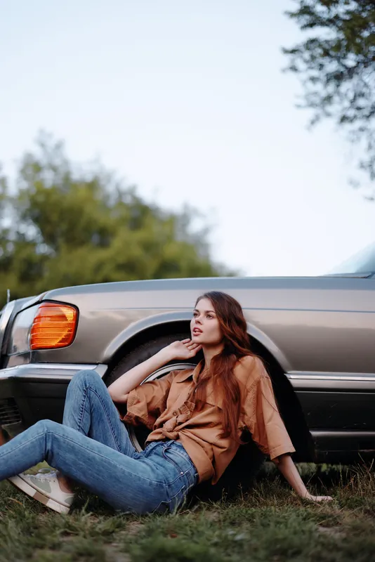 Young woman in casual attire sitting beside a vintage car, expressing relaxed vibes amidst nature, with a soft color palette and seasonal autumn backdrop.