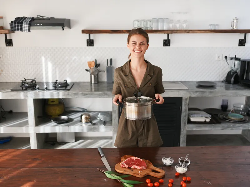 Женщина на кухне с кастрюлей в руках улыбается, готовит ужин из мяса и овощей на деревянном столе на фоне стильной кухни, образ жизни