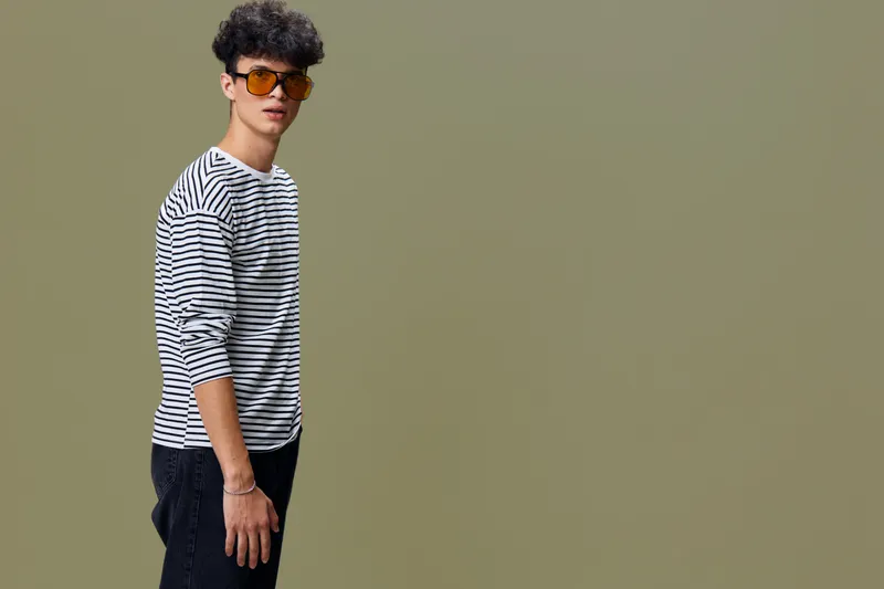 Young man wearing sunglasses and a striped shirt standing against an olive green background, showcasing casual style and modern fashion in a studio setting.