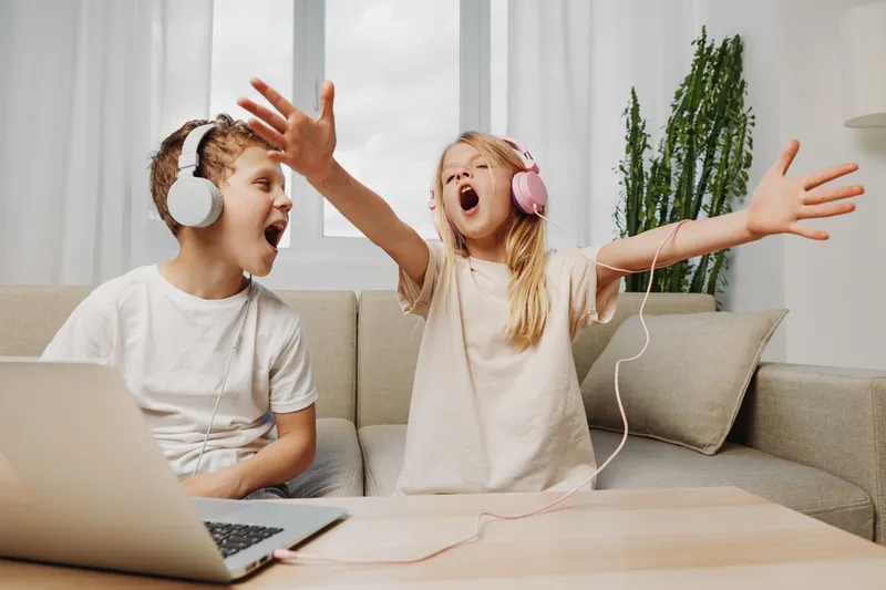 Two children wearing headphones enjoy music and sing passionately while sitting on a sofa with a laptop in a bright living room environment.