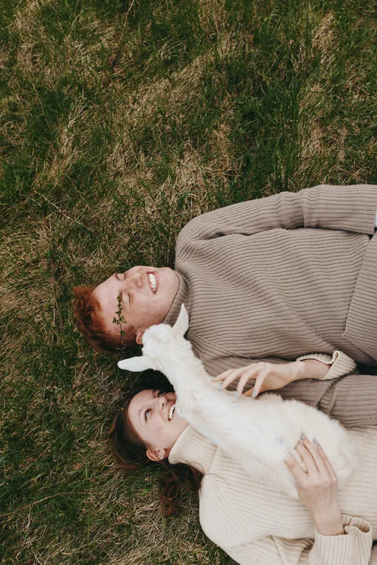 Lifestyle moment of a couple outdoors on grass with a playful goat kid, cozy sweaters, warm light, candid affection, peaceful atmosphere, natural colors, gentle companionship, rustic charm