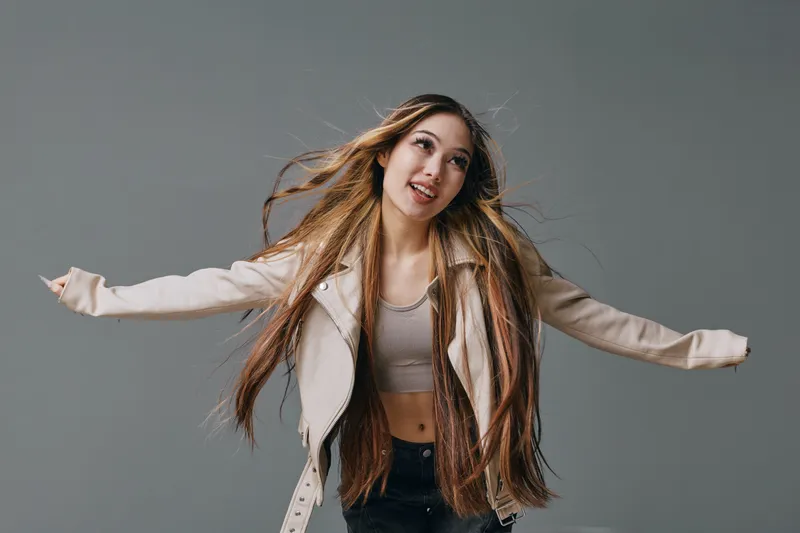 Young woman with long braided hair wearing a light jacket and crop top posing with arms outstretched isolated on grey background. Casual fashion, hairstyle and expression.