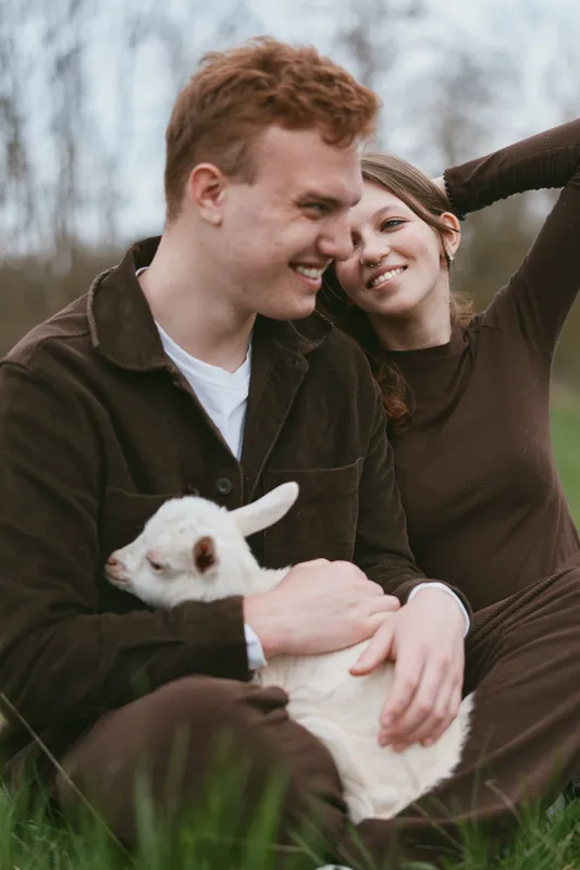 Young couple sits on grass holding a white baby goat while enjoying a relaxed moment outdoors in casual brown clothing during a calm overcast day.
