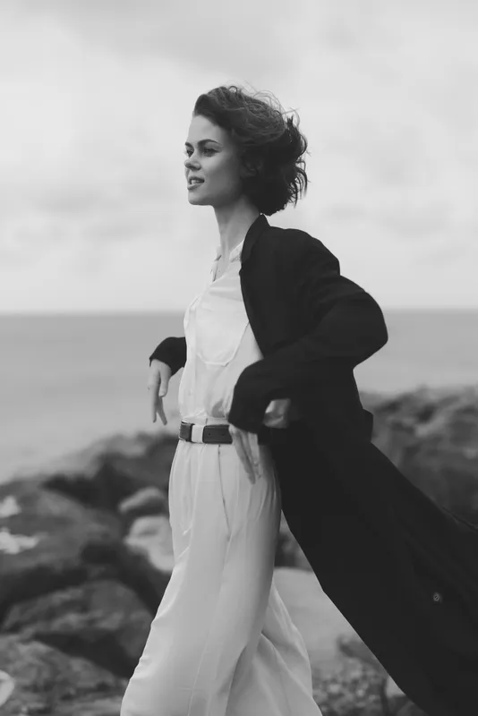 Woman walks along a windswept coastline in a flowing coat, monochrome scene, conveying movement, fashion sense, and seaside mood.