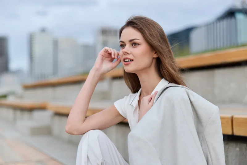Woman in light beige outfit sitting outdoors with thoughtful expression on urban background, fashion style, natural light, modern cityscape