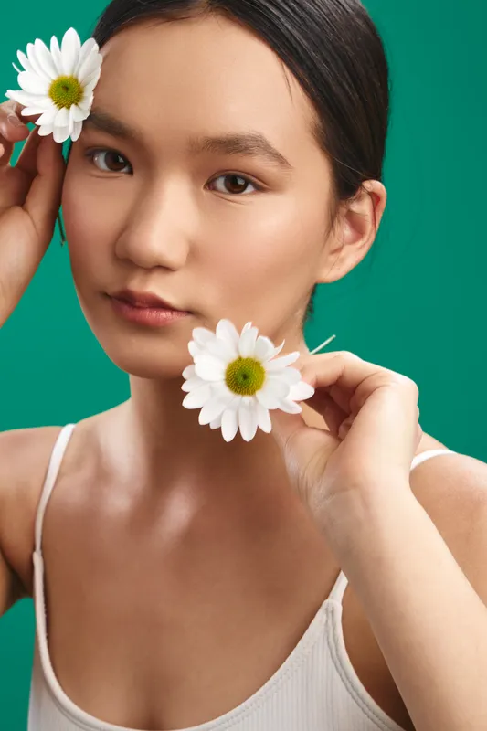 Asian woman with a delicate smile holds daisies near her face in a clean studio portrait, conveying fresh beauty and serene charm against a vibrant backdrop.