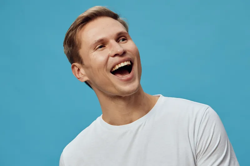 Smiling happy young man with light skin and short brown hair in white t-shirt looks joyful against a bright turquoise background studio portrait. People lifestyle concept, positive emotion, relaxed mo