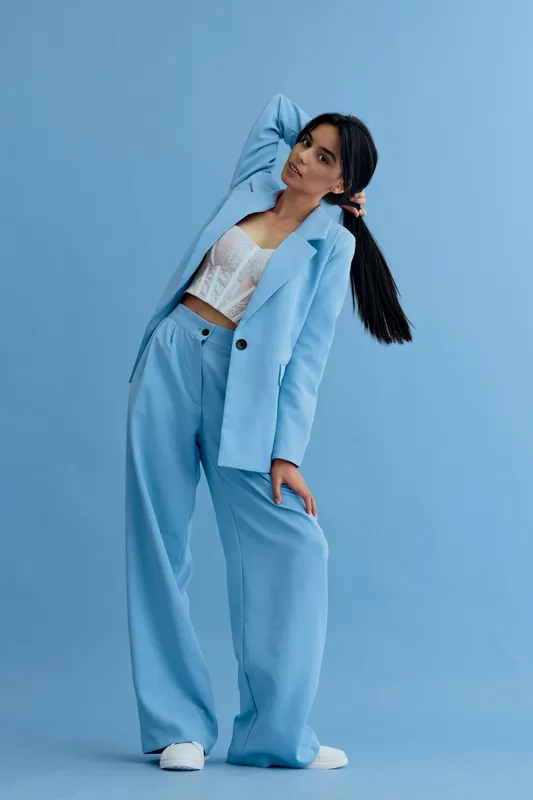 young woman in blue pantsuit poses confidently against a solid blue background, wearing a white lace top and white sneakers, modern fashion style portrait.