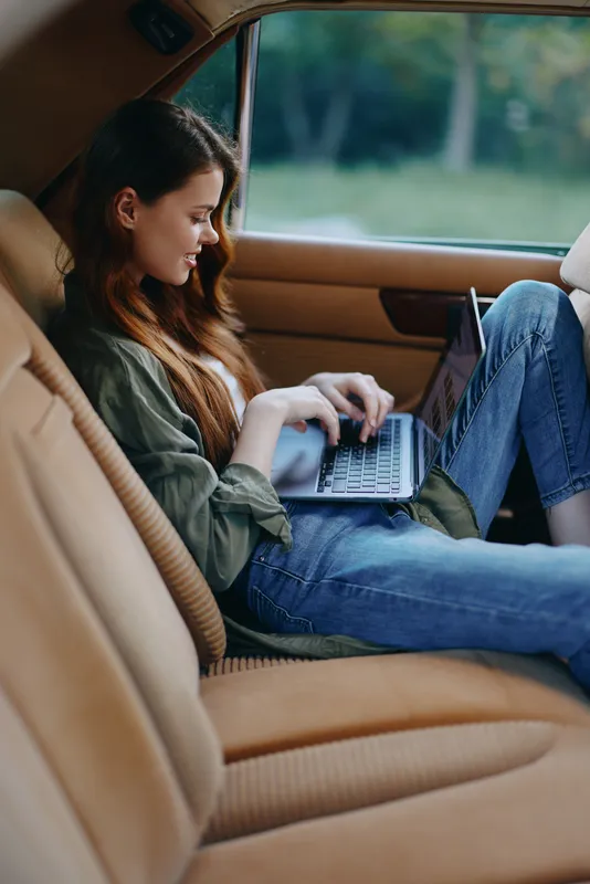 Young woman working on a laptop in a car, showcasing digital nomad lifestyle, casual style, soft natural light, and autumn vibes.
