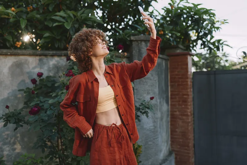 A stylish woman in a rust red jacket poses outdoors, gazing upward with a bright smile. A simple garden backdrop highlights casual fashion and confident mood